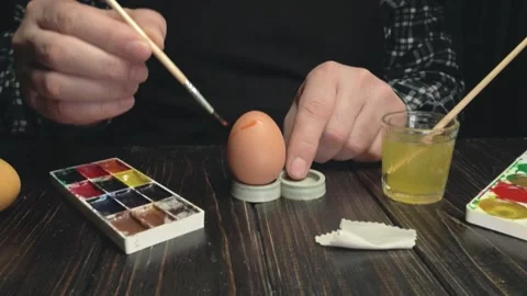 Person painting an egg with a brush, using watercolor palette and glass of Stock Footage 337904575