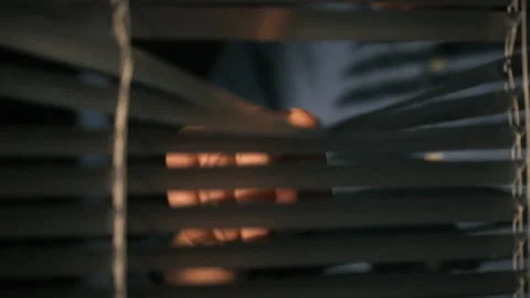 Person peering through blinds in a dimly lit room during nighttime hours Stock Footage 323869915