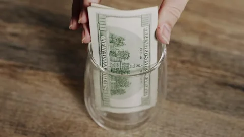 Person Placing Stack of One Hundred Dollar Bills into Glass for Savings Concept 動画素材 340857206