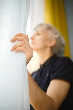 Person pulls back curtain to look outside during morning light in indoor spac Stock Photos