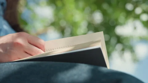 Person reading book on lap under tree slow page turns denim lap dappled sunlight Stock Footage 329477969