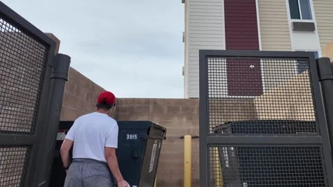 Person in red cap tosses white trash bag into garbage dumpster Stock Footage 340429823