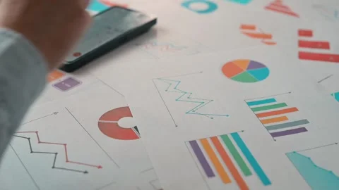 A person is reviewing various graphs and charts laid out on a table while using Stock-Footage 338302453