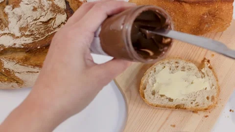 Person Scooping Chocolate Hazelnut Spread from Jar onto Sliced Bread Stock Footage 337395055