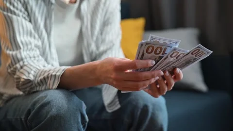 A person is seated on a couch counting a stack of cash in a comfortable indoor Video stock 338446136