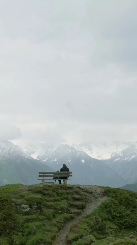 A person sits on a bench overlooking snow-capped mountains in the Tirol region 動画素材 285472012