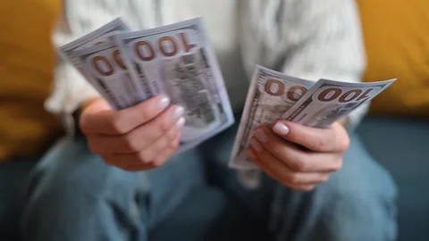 A person sits on a couch and counts a stack of cash with bills visible in their Stock-Footage 338446163