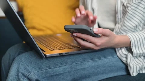 A person sits on a couch, using a smartphone while working on a laptop placed on Stock Footage 338446294