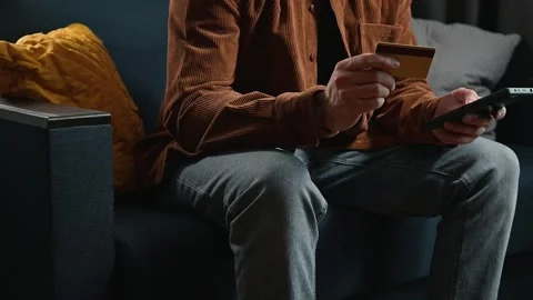 A person sits on a couch while holding a credit card and a remote in a living Stock-Footage 339097685