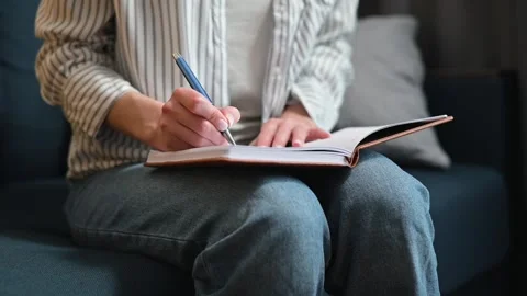 A person sits on a couch, writing in a notebook with a pen. The individual Stock Footage 338446357