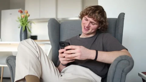 A person sits happily, smiling while looking at his smartphone. The cozy chair,  Stock Footage 302633829