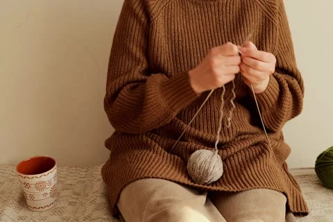 A person sits on a light surface and knits using a ball of yarn. A cup sits Stock Footage 336464917