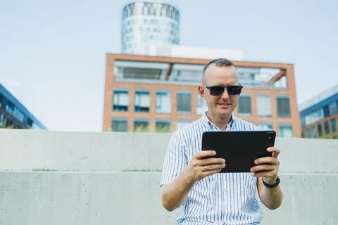 Person sits on outdoor steps using a tablet in an urban area with modern bu.. Stock Photos