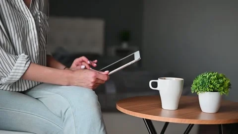 A person sits at a small table holding a tablet. A cup of coffee and a small Stockbeeldmateriaal 336778522