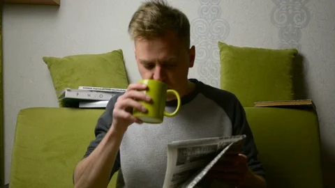 Person sits on a sofa drinking tea and starts to read an old newspaper 스톡 동영상 92947376