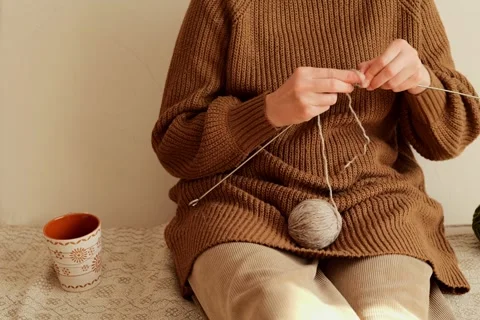 A person sits on a soft surface while knitting, holding needles and yarn, with a Видео 336464919