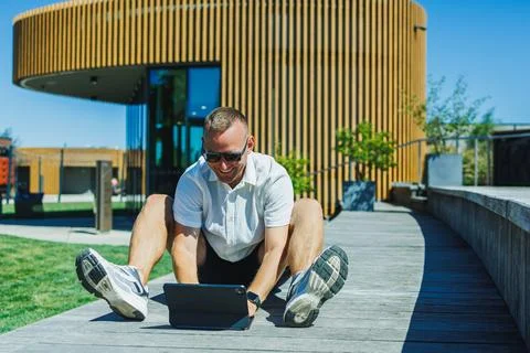 Person sits on wooden deck using tablet in outdoor area near a modern build.. Stock Photos