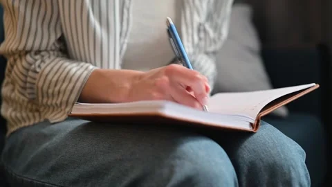 A person is sitting comfortably while writing in a notebook using a blue pen. Stock Footage 338446381