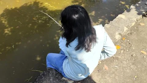 A person sitting by a pond, looking down at the water. The scene captures a.. Fotos Stock