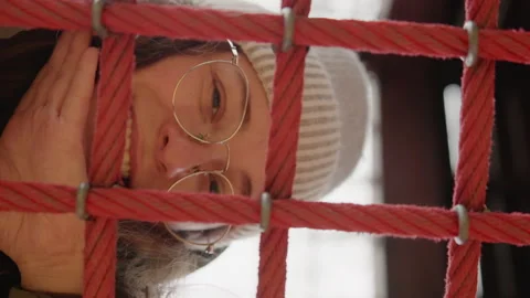 Person smiling through red rope grid, eyeglasses and knit beanie, playful peek Stock Footage 345539790
