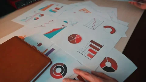 A person is sorting through various printed graphs and charts on a desk. The Stock-Footage 338302799