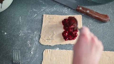 A person spreads jam onto a sheet of dough in a kitchen setting, using utensils Stock Footage 339095664