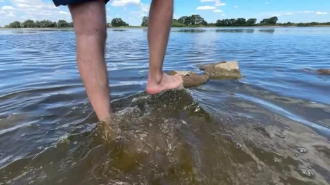 Person standing on a stone in a river, camera pans up to reveal scenic river vie Stock Footage 339315188