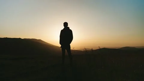 Person stands on a hill at sunset looking towards the horizon with mountains in  Stock Footage 343843221