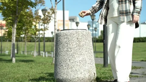 Person throwing waste into stone trash bin in sunny urban park with trees and Stock Footage 345527998