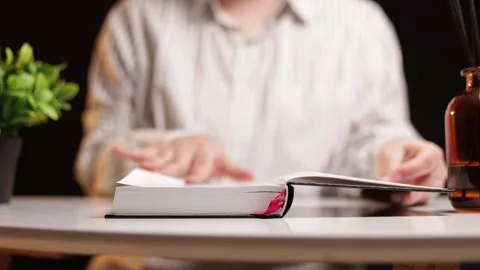 Person turning pages of a book while sitting at a table with a plant and drink Stock Footage 339215714