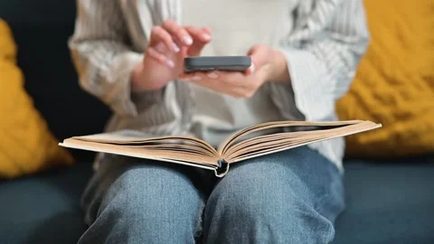 A person turns the pages of a book while interacting with a smartphone in a cozy Stock Footage 338446497