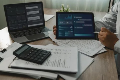 Person uses AI assistant interfaces on a laptop and tablet above paperwork .. Stockfoto's