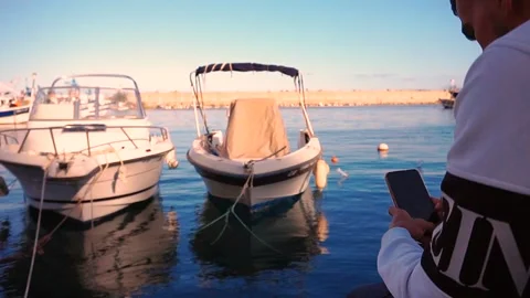 A person uses his phone while sitting in the port with ships in front of him 스톡 동영상 165243381