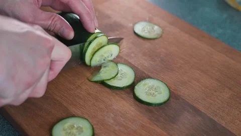 A person uses a knife to slice a cucumber on a wooden cutting board in a kitchen Stock-Footage 338290623