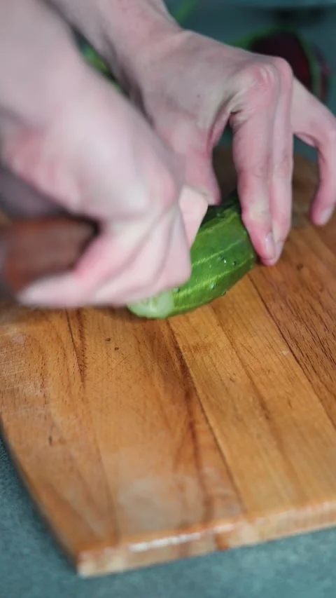 A person uses a knife to slice a cucumber on a wooden cutting board. The hands Stock Footage 338304167