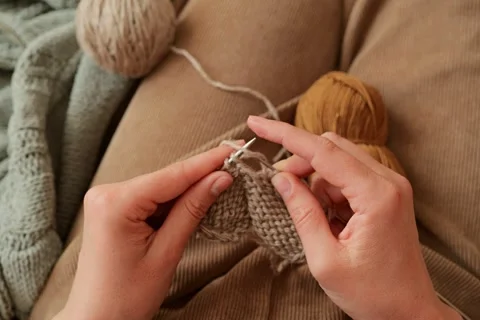 A person uses knitting needles to create a piece of fabric while sitting on a 스톡 동영상 336464836