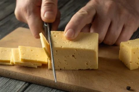 A person uses scissors to cut a block of yellow cheese into slices on a woo.. Stock Photos