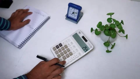 Person using a calculator on a desk with a notebook, clock, and plant Stock Footage 317131652