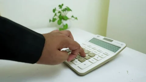 Person using a calculator on a white desk with a plant in the background Stock Footage 320725591