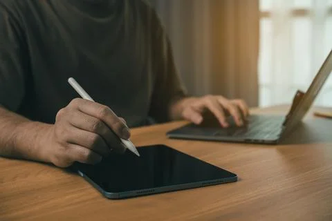 Person using digital pen on tablet screen while working on laptop at wooden.. Stock Photos