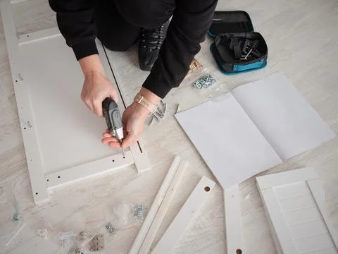 A person using an electric screwdriver to assemble white wooden furniture. Stock Photos
