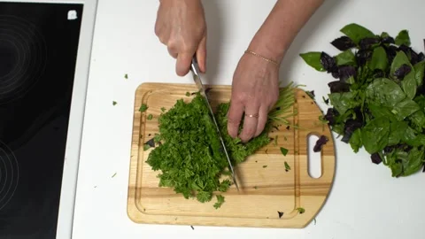 A person is using a knife to finely chop green herbs on a wooden cutting board, Video stock 336443204