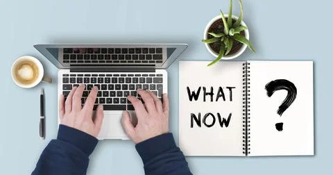 Person using laptop computer besides notepad with text WHAT NOW work planning Stock Photos