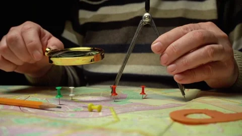 Person using a magnifying glass and compass to analyze a map with colorful push Stock Footage 332940331