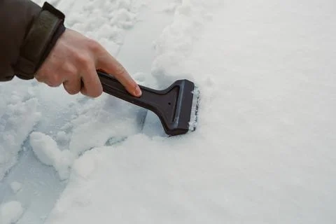 A person is using a scraper to clear snow off the roof of a car. The ground i Stock-Fotos