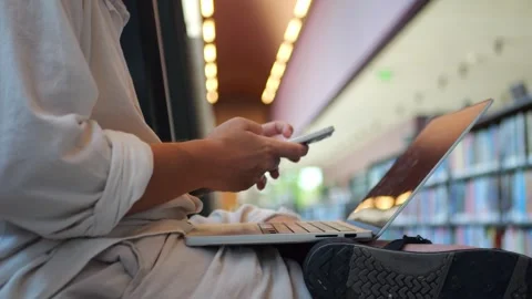 Person Using Smartphone Beside Laptop in Library, Modern Multitasking... Vidéo 334437049