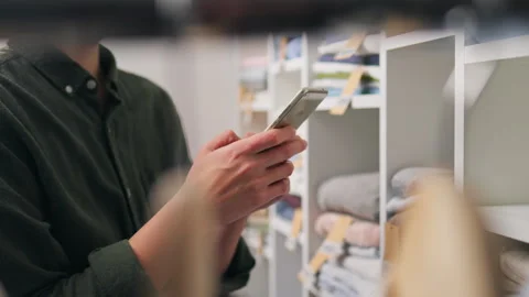 Person using smartphone while shopping for clothes in a neatly organized ret Stock Footage 323714191