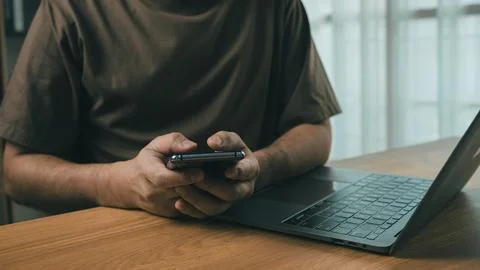 Person using smartphone while sitting at wooden desk with open laptop in mo.. Stock Photos
