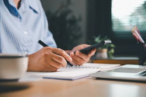 Person using a smartphone while writing notes beside a laptop, representing.. Stock Photos