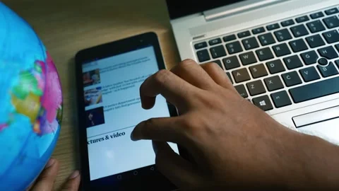 Person using a tablet next to a laptop and globe on a wooden desk Stock Footage 317736763